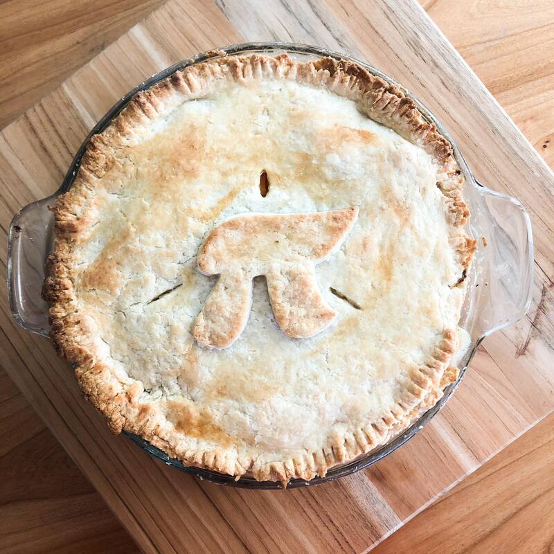 A homemade pie in a glass pie dish has the mathematical symbol for pi shaped with dough on the top crust.  