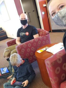 Becca holds the phone for a selfie. Robert is sitting in a waiting room chair and smiling behind a mask. 2 boys sit on the floor playing video games on tablets. 