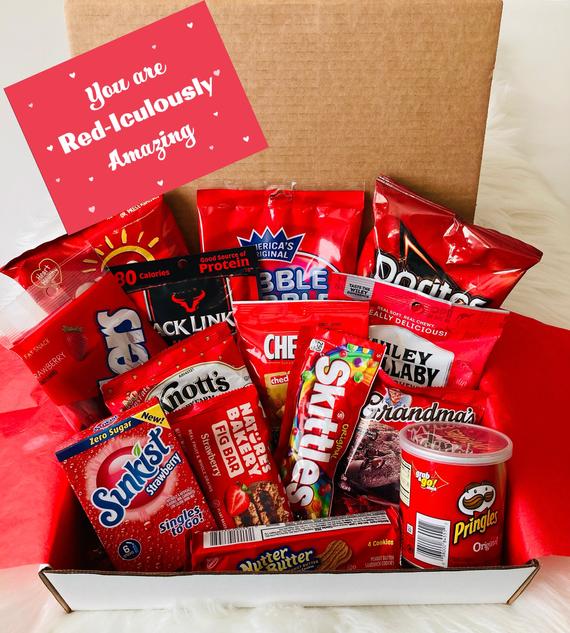 A box is open showing many different snacks with red packaging: chips, candy, cookies, beef jerky, granola bars. They are displayed on tissue paper. The text reads 