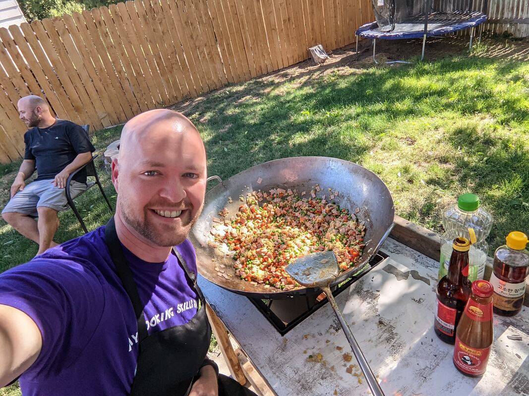 Bald caucasian man in apron smiles at a selfie with a large wok full of food behind him and sauces on the counter. Grass and fence are visible behind him in the yard.